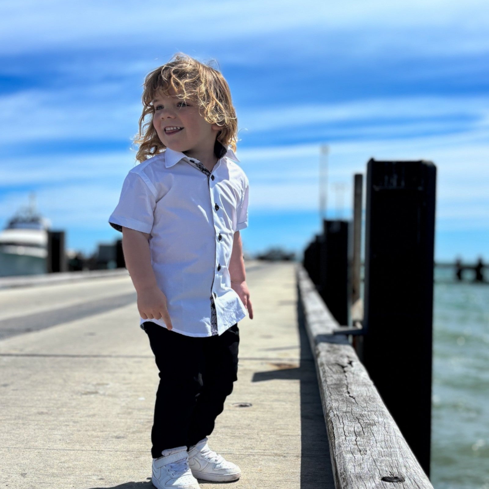 Child standing on a wooden dock by the water with a clear blue sky.